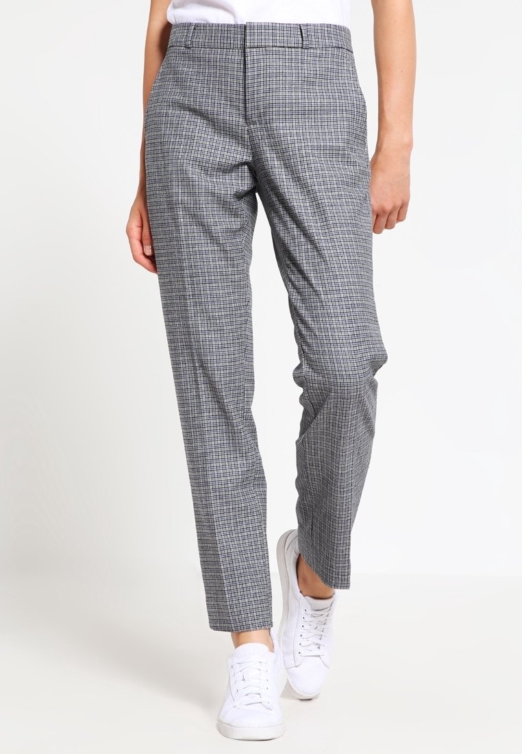 Kvinna i grå rutiga slim-fit byxor och vita sneakers, stående mot en enkel vit bakgrund.