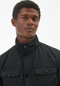 Zwarte jas met een hoge kraag, drukknoopsluiting en twee borstzakken. De stof heeft een gladde textuur en een strakke, minimalistische uitstraling.