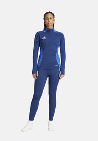 Marinefarbener Sportoberteil mit blauen Akzenten, langen Ärmeln, hoch geschlossenem Kragen und Reißverschluss; kombiniert mit passenden Leggings und weißen Sportschuhen.