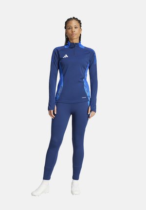 Marinefarbener Sportoberteil mit blauen Akzenten, langen Ärmeln, hoch geschlossenem Kragen und Reißverschluss; kombiniert mit passenden Leggings und weißen Sportschuhen.