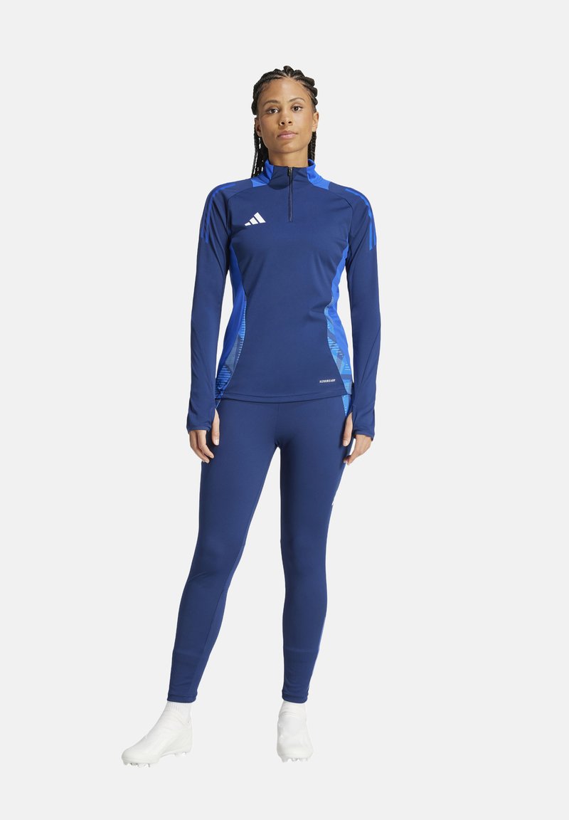 Marinefarbener Sportoberteil mit blauen Akzenten, langen Ärmeln, hoch geschlossenem Kragen und Reißverschluss; kombiniert mit passenden Leggings und weißen Sportschuhen.