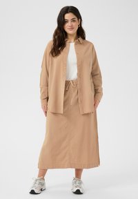 Camisa de botones marrón con mangas largas sobre una falda de cordón a juego. Ambos son de una tela suave, con un ajuste relajado, combinados con zapatillas.