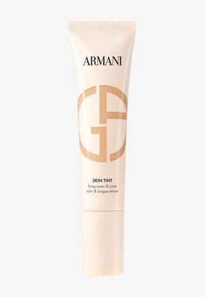 Tubo de color beige claro con un acabado brillante y un degradado de blanco a dorado, que presenta "ARMANI" y un logotipo circular. El texto indica "SKIN TINT de larga duración y cuidado."