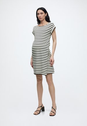 Zwangere vrouw die een zwart-wit gestreepte knie lengte jurk en zwarte sandaaltjes met bandjes draagt, staand tegen een effen witte achtergrond.