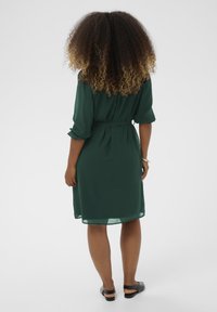 Roheline, voolav kleit vöö ja kolmeveerandvarrukatega. Läbipaistev kangas, millel on sile tekstuur. Vaade seljalt, paljastades lihtsa disaini.