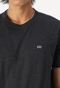 Personne portant un t-shirt gris foncé avec un petit logo blanc "GAP" sur la poitrine gauche et une chaîne argentée autour du cou.