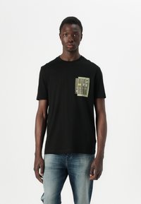 T-shirt nera in cotone a maniche corte, con un taschino verde decorato da grafiche stampate in forma rettangolare sul lato sinistro del petto.