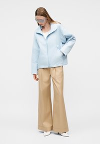 Femeie purtând o geacă supradimensionată albastru deschis, pantaloni largi bej, pantofi albi cu toc ascuțit și ochelari de protecție transparenți, stând cu o mână în buzunar.