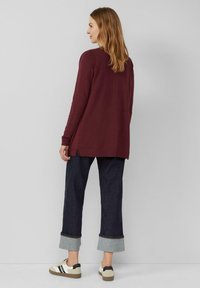 Burgunderfarbenes Langarmoberteil mit seitlichen Schlitzern, kombiniert mit dunklen Jeans im Denim-Stil mit umgeschlagenem Saum und beigen Sneakers mit schwarzen Akzenten.