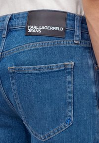 Гледка отзад на сини дънки с черен етикет с надпис Karl Lagerfeld Jeans над задния джоб.