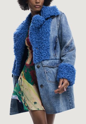 Femme portant un manteau en denim bleu avec un col et des poignets en fourrure bouclée bleu vif, par-dessus une robe à motif abstrait coloré.