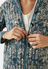 Chaqueta acolchada en azul-verde con patrón floral, presenta dos bolsillos frontales y un ribete en contraste. Se cierra con lazos y toggles.