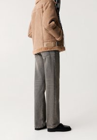 Veste en shearling beige avec un col doux et des poches latérales, associée à un jean gris ample et des chaussures noires. Lignes épurées et design minimaliste.