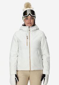 Weiße isolierte Schneejacke mit einem Frontreißverschluss, braunen Akzenten und einer Kapuze. Dazu beige Skihose, Handschuhe und eine gestrickte Mütze mit Bommel.