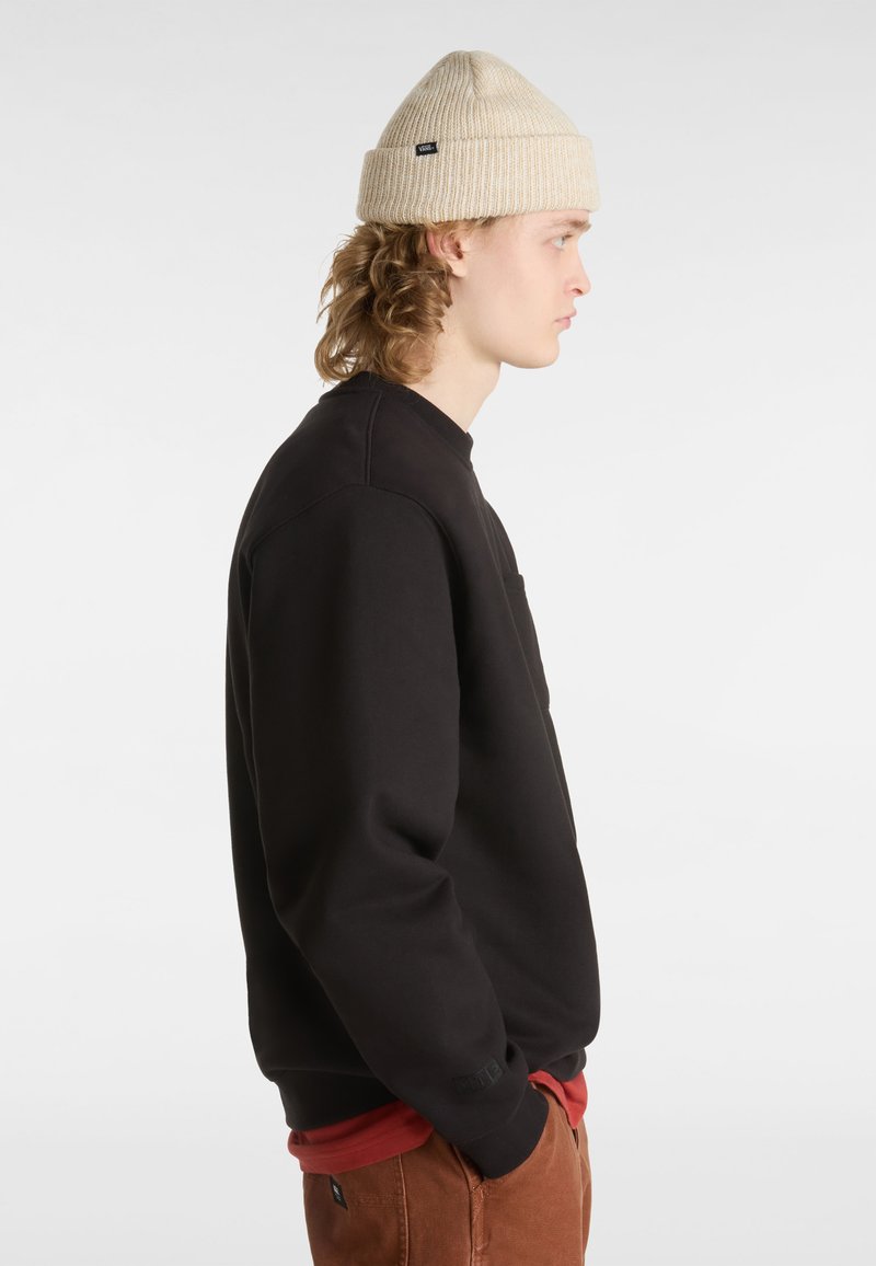 Zwarte sweatshirt met een ronde hals, zijzak en geribbelde manchetten, gecombineerd met een lichtbeige gebreide beanie. Model in profiel weergegeven.