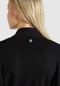 Femme blonde portant un pull en maille noire avec un petit emblème métallique rond en haut du dos, vue de dos sur un fond uni.