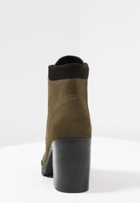 Olijfgroen suède enkelboot met een zwarte gladde manchette en een dikke zwarte hak. Het ontwerp heeft een ronde neus en minimale details.