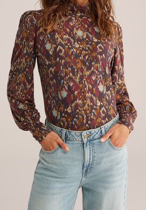 Vrouw met een blouse met lange mouwen en een hoge nek, bedrukt met een meerkleurige abstracte print, en lichtblauwe jeans met hoge taille, handen in de zakken.