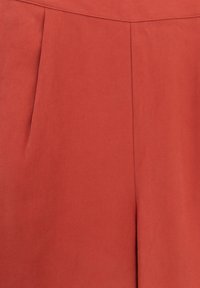 Un pantalon couleur rouille en tissu lisse et texturé présente des plis marqués et un design de taille épuré. Aucun matériel ou accent visible.