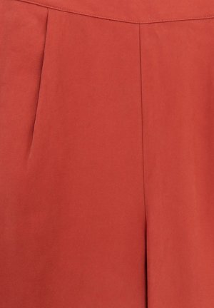 Un pantalon couleur rouille en tissu lisse et texturé présente des plis marqués et un design de taille épuré. Aucun matériel ou accent visible.