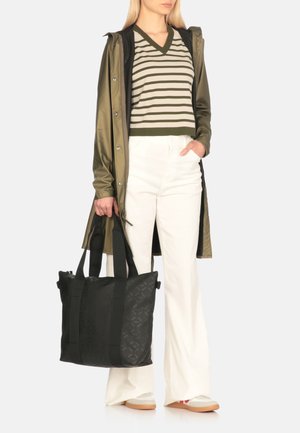 Borsa a tracolla nera con motivo testurizzato, tenuta da una persona che indossa un maglione a righe verdi e un impermeabile oliva sopra pantaloni bianchi a zampa d'elefante.