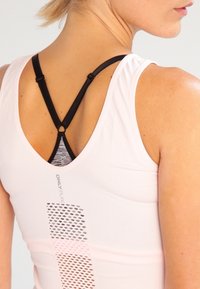 Ljusrosa träningslinne med V-ringning, svarta justerbara axelband och en meshpanel med ett geometriskt mönster på baksidan.