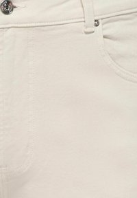 Prim-plan al unei perechi de blugi din denim alb, care prezintă nasture metalic, nit, buclă pentru curea și buzunar frontal cu cusături vizibile.