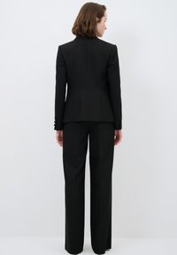 Musta räätälöity puku, jossa on istuva bleiseri ja suorat housut. Sujuva kangas, jossa on minimaalista yksityiskohtaisuutta, napit hihoissa ja jämäkkä siluetti.