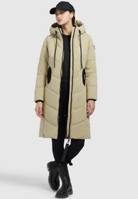 Gaišbēša polsterēta garā jaka ar kapuci, melni akcentu pleķi, priekšējais rāvējslēdzis un auklas, apvienota ar melnām legingām un zābakiem.