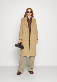 Beige Wollmantel mit Reverskragen und Seitentaschen, kombiniert mit einem braunen Hemd und beigen Palazzo-Hosen. Schwarze Handtasche.