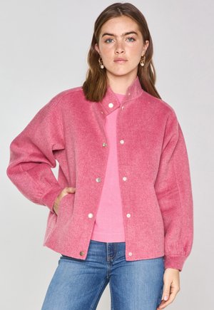 Veste en laine mélangée floue et rose avec des boutons-pression argentés, un design sans col et des poches latérales. Portée sur un haut rose et associée à un jean bleu.