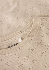 Jasnobeżowy sweter dziergany na drutach z motywem kokardki, okrągłym dekoltem i płaskim przeszyciem oraz metką "name it".