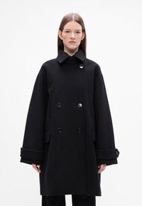 Cappotto di lana nera con colletto alto, doppio petto e grandi tasche. Presenta maniche ampie e un design strutturato sottile.