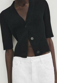 Schwarze gerippte Strickjacke mit V-Ausschnitt, kurzen Ärmel und zwei dunklen Knöpfen, kombiniert mit weißen Jeans.