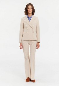 Blazer beige croisé avec des boutons ronds, associé à un pantalon assorti avec un motif texturé, porté sur une chemise bleue. Mocassins marron.