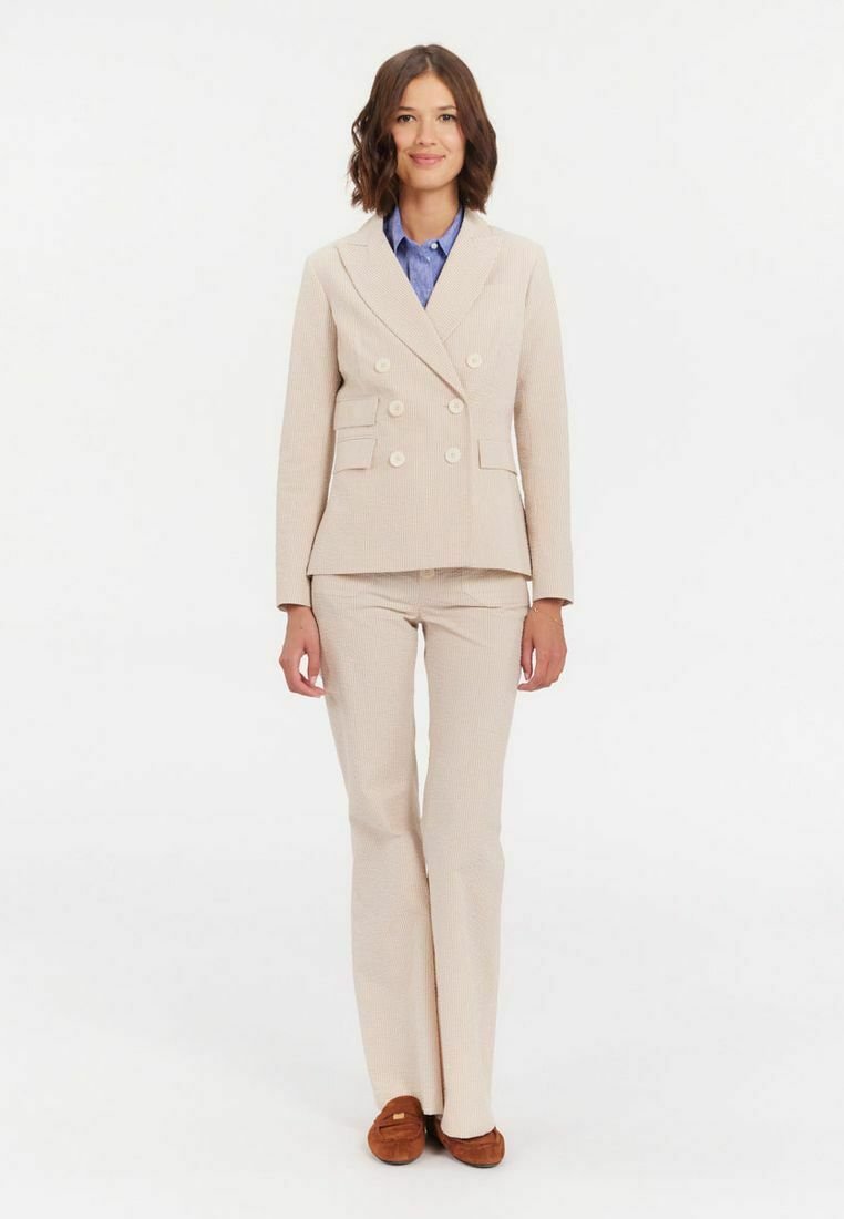 Blazer beige croisé avec des boutons ronds, associé à un pantalon assorti avec un motif texturé, porté sur une chemise bleue. Mocassins marron.