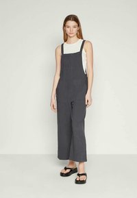 Mala cinza escura com um corte largo, alças ajustáveis, grandes bolsos frontais e um tecido com textura suave, combinada com uma blusa branca e sandálias.