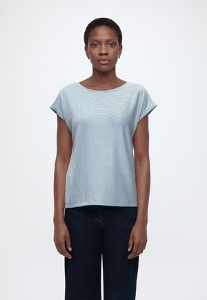 Kobieta o krótkich włosach, ubrana w jasnoniebieską koszulkę w paski na krótki rękaw i ciemne spodnie, stojąca na białym tle.