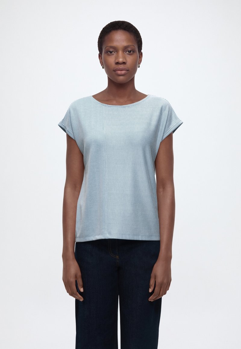 Vrouw met kort haar, gekleed in een lichtblauw gestreept topje met korte mouwen en een donkere broek, staand tegen een witte achtergrond.