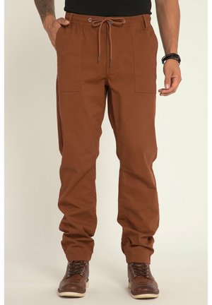 Uomo che indossa pantaloni marroni con coulisse e tasche applicate frontali e stivali marroni in pelle con lacci su sfondo chiaro.