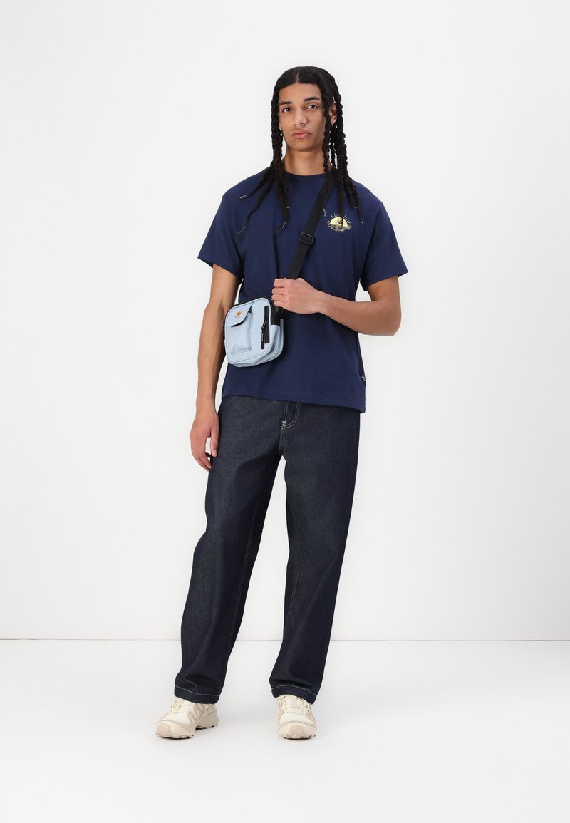 Camiseta azul marino con un pequeño emblema, jeans de mezclilla oscura de corte relajado, bolso cruzado azul claro con un bolsillo frontal y zapatillas crema.
