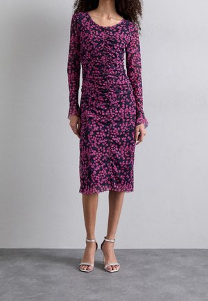 Kvinde iført en langærmet, knælængde, mørk kjole med pink blomsterprint og rynket forstykke, kombineret med sølvfarvede højhælede sandaler.