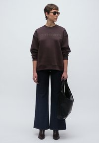 Sötétbarna pulóver "DOLCE FAR NIENTE" felirattal, széles szárú sötét farmerrel, fekete válltáskával és barna, magas sarkú csizmával párosítva.