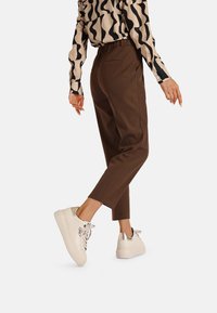 Pantaloni marroni a sigaretta con vita alta, abbinati a sneakers bianche con dettagli argentati. Camicia a maniche lunghe a righe nere e beige.