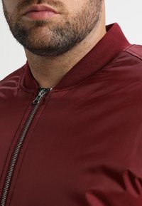 Burgunderfarget bomberjakke med et glatt, skinnende stoff, med glidelås, ribbet krage og subtil tekstur. Nærbilde av overkroppen.
