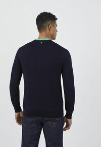 Maglione lavorato a maglia blu navy con maniche lunghe, scollatura rotonda e un accento verde sul colletto interno. Presenta un dettaglio con logo sul retro.