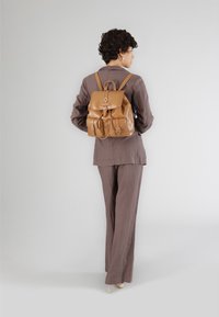 Läderryggsäck i brunt med lockstängning, två framfickor och justerbara axelremmar. Modellen bär en brun kostym.