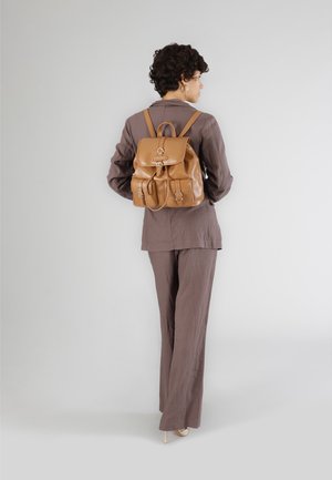 Sac à dos en cuir tan avec une fermeture rabat, deux poches avant et des bretelles réglables. Le modèle porte un costume marron.