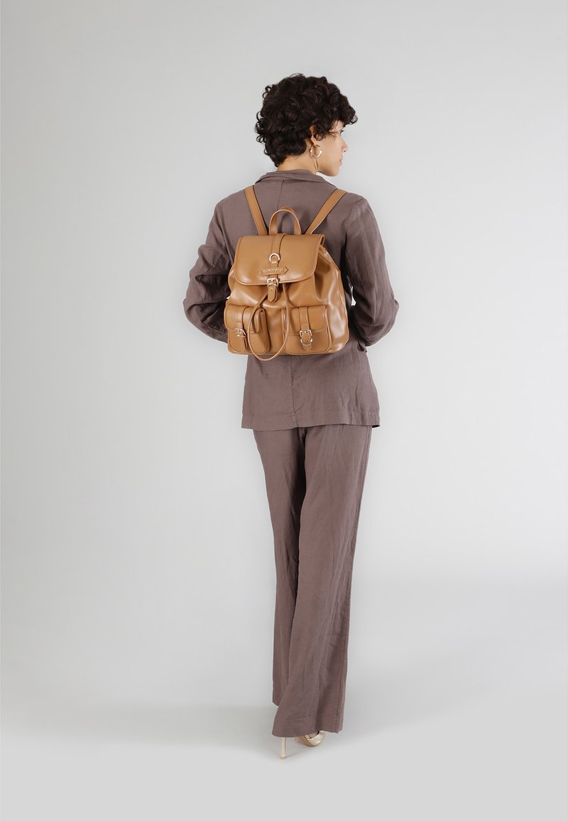 Läderryggsäck i brunt med lockstängning, två framfickor och justerbara axelremmar. Modellen bär en brun kostym.