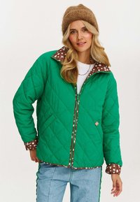 Zielona pikowana kurtka z brązowym kołnierzem w kropki i mankietami, z zamkiem błyskawicznym z przodu i luźnym krojem. Dopełniona niebieskimi dżinsami.
